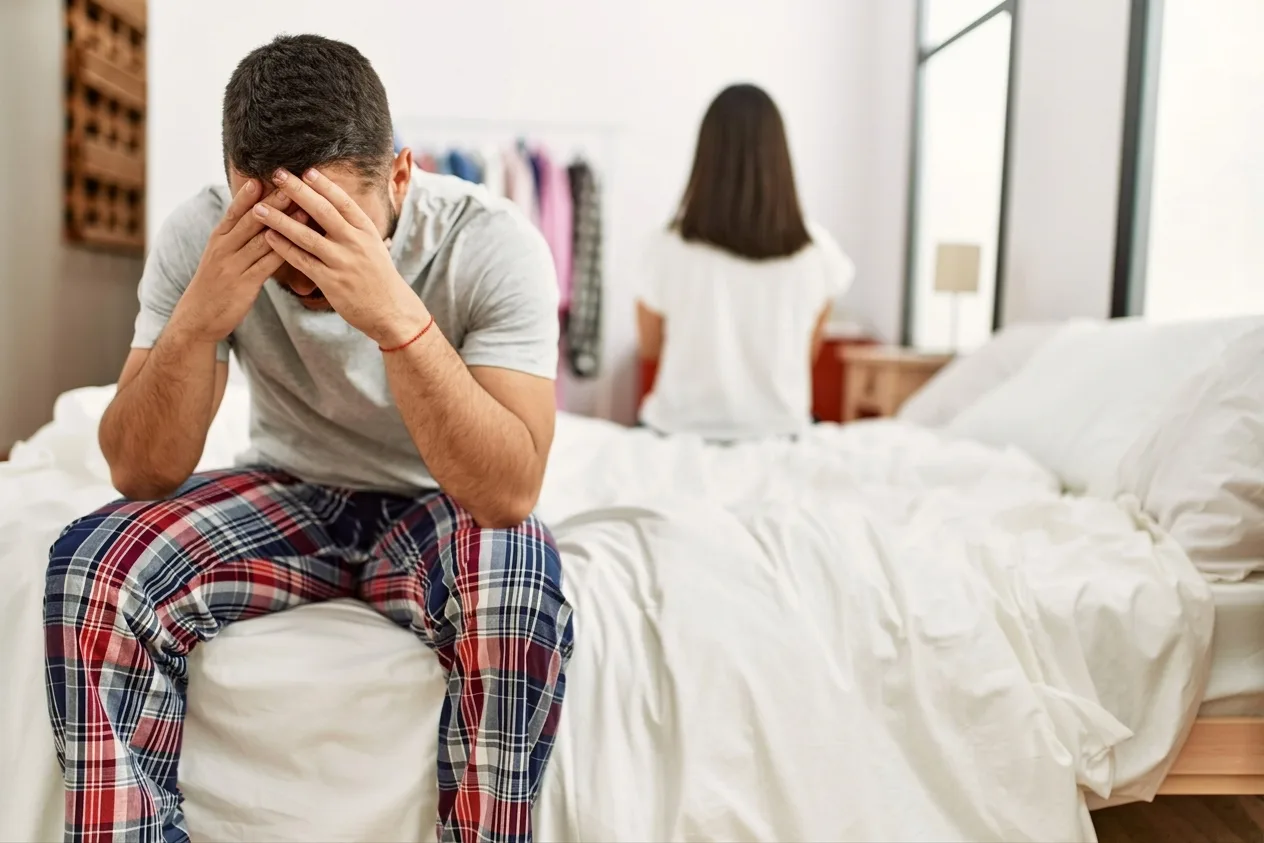 Man sitting on the edge of the bed with his head in his hands, reflecting the strain of sexual dysfunction